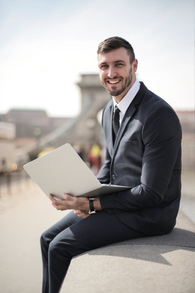 Homme d'affaires heureux, en costume, tenant son ordinateur portable, assis sur un pont de la ville, illustrant le style de vie moderne des entreprises.