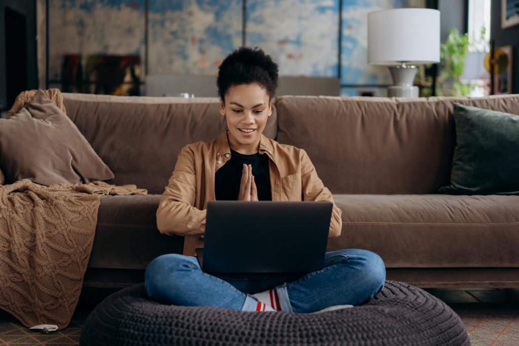 Une jeune femme souriante est assise sur un pouf, les jambes croisées, et utilise son ordinateur portable dans un cadre domestique confortable.