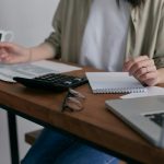 Une femme gère ses finances à la maison, à l'aide d'un ordinateur portable et d'une calculatrice posés sur un bureau en bois.