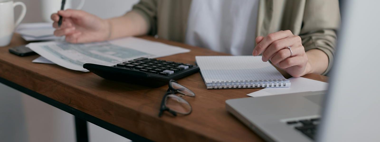 Une femme gère ses finances à la maison, à l'aide d'un ordinateur portable et d'une calculatrice posés sur un bureau en bois.