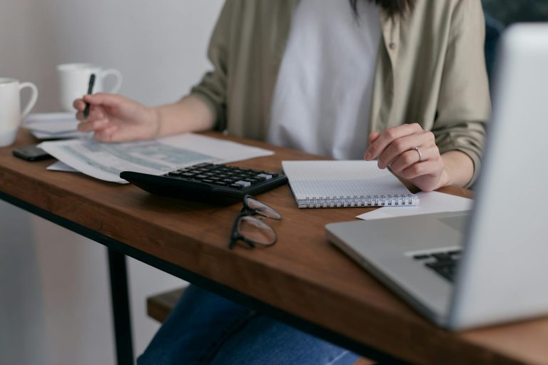 Une femme gère ses finances à la maison, à l'aide d'un ordinateur portable et d'une calculatrice posés sur un bureau en bois.